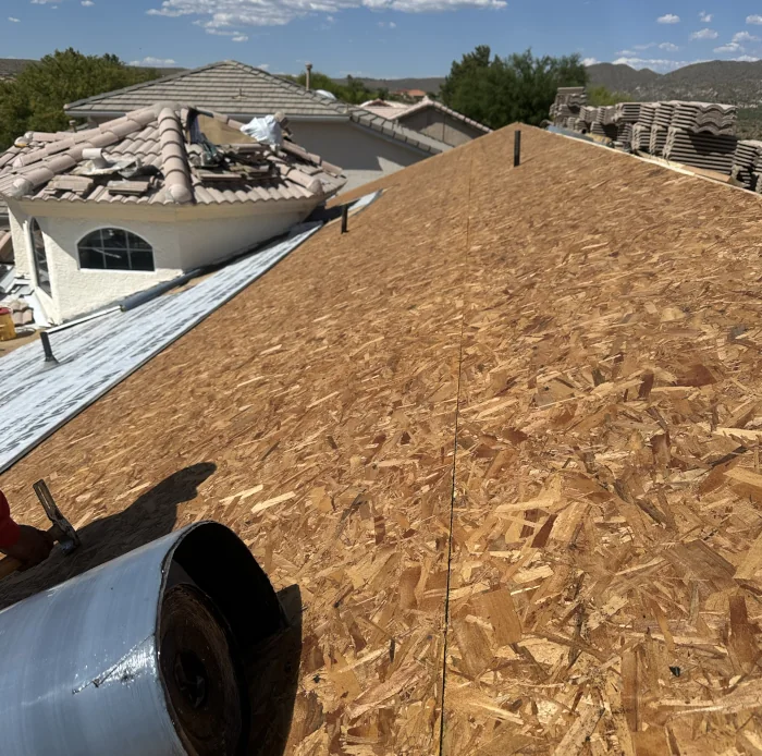 Wide view from a rooftop showing a newly sheathed pitched roof of oriented strand board (OSB) with a roll of roofing material and stacks of tiles nearby; neighboring homes with tile roofs and distant hills are visible under a partly cloudy sky.
