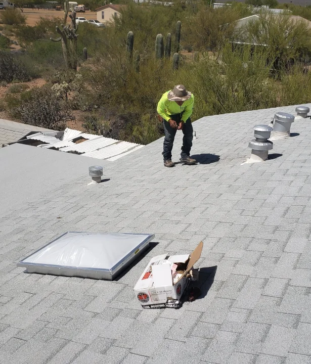 Worker in a bright yellow‑green shirt and hat on a light-gray shingled roof beside vents, a skylight, and an open box; desert vegetation with cacti visible beyond.